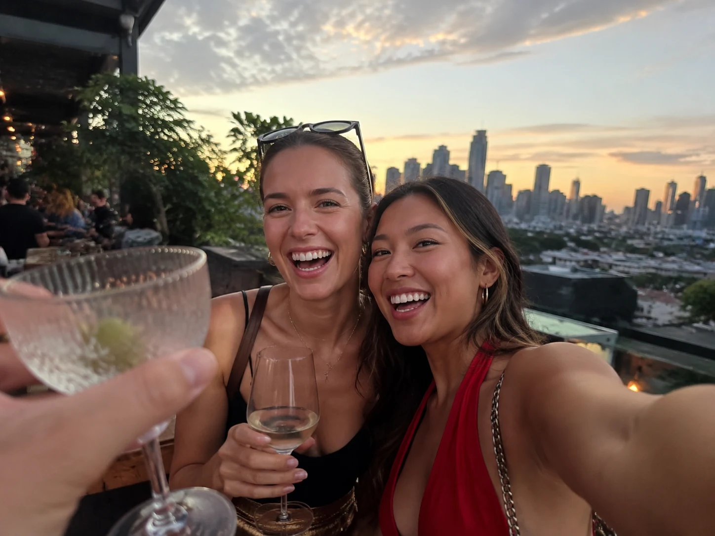 Two friends taking a rooftop bar selfie with a stranger's hand and cocktail glass intruding from the left edge of the frame