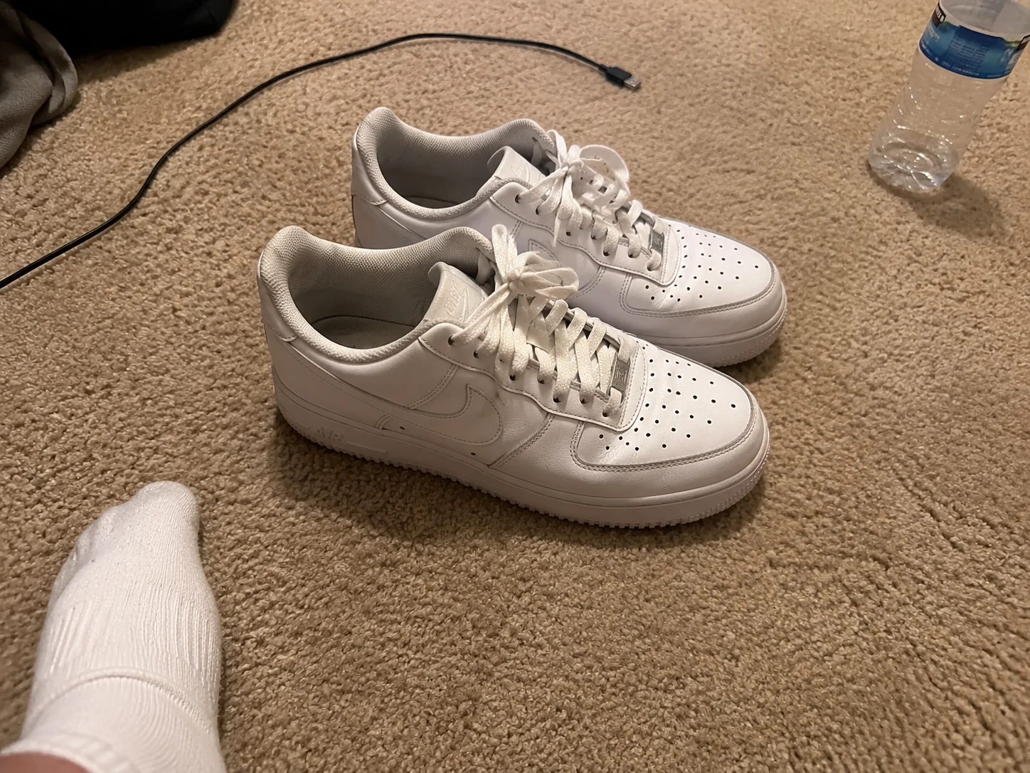 White Nike Air Force 1 sneakers on carpet with the photographer's foot and sock, a charging cable, and a water bottle visible in the frame