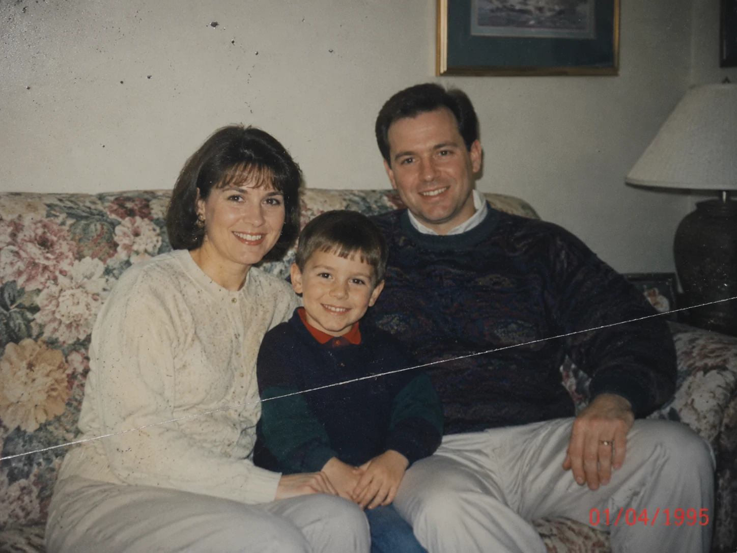 Scanned 1995 family photo of parents and young son on a floral sofa, showing a diagonal scratch across the lower half, dust spots, and a red date stamp