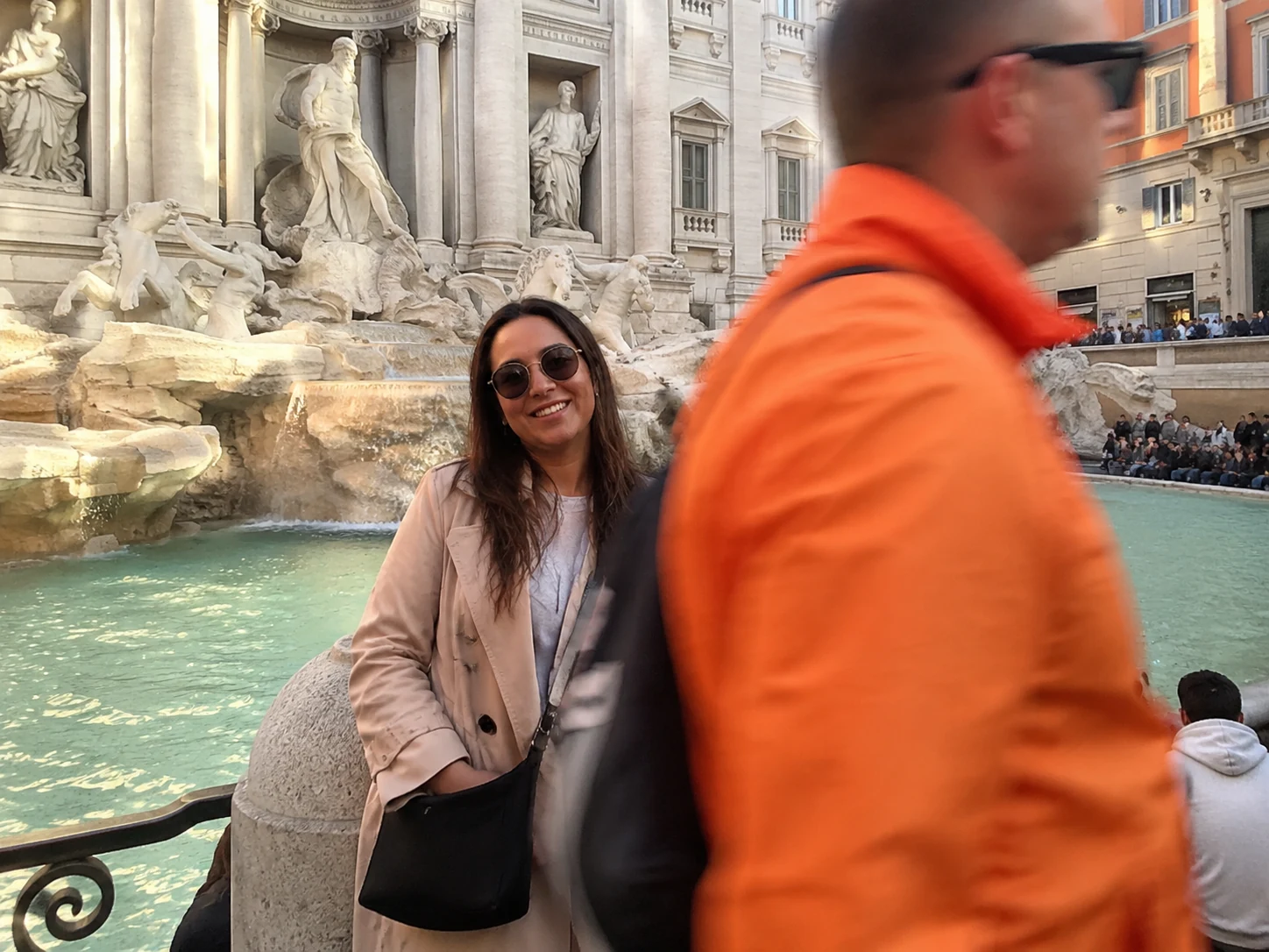 Woman posing at the Trevi Fountain in Rome with a tourist in an orange jacket walking through the foreground blocking the shot